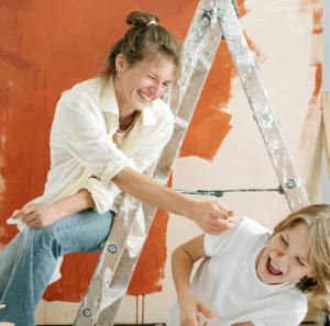 Femme et enfant souriants en train de peindre un mur avec une couleur orange dans un salon en rénovation.