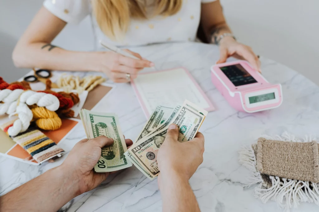 Deux personnes comptant de l'argent en billets de 20 et 100 dollars, avec des éléments créatifs sur une table.