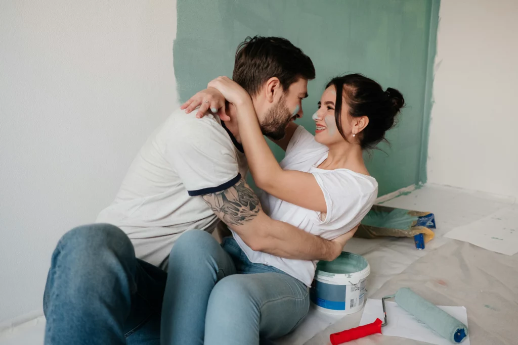 Couple souriant en pleine rénovation de salon, assis près d’un mur partiellement peint en vert clair avec des outils de peinture à leurs côtés.