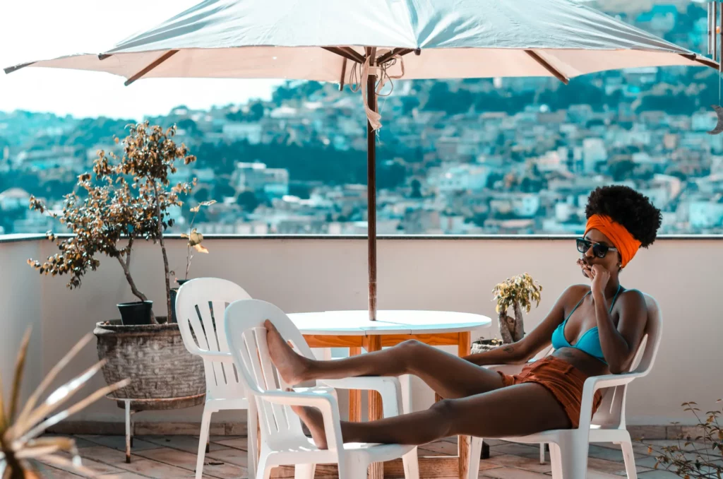 Femme détendue en maillot de bain et short, assise sur une terrasse avec parasol, entourée de chaises blanches et plantes en pots, vue urbaine en arrière-plan.
