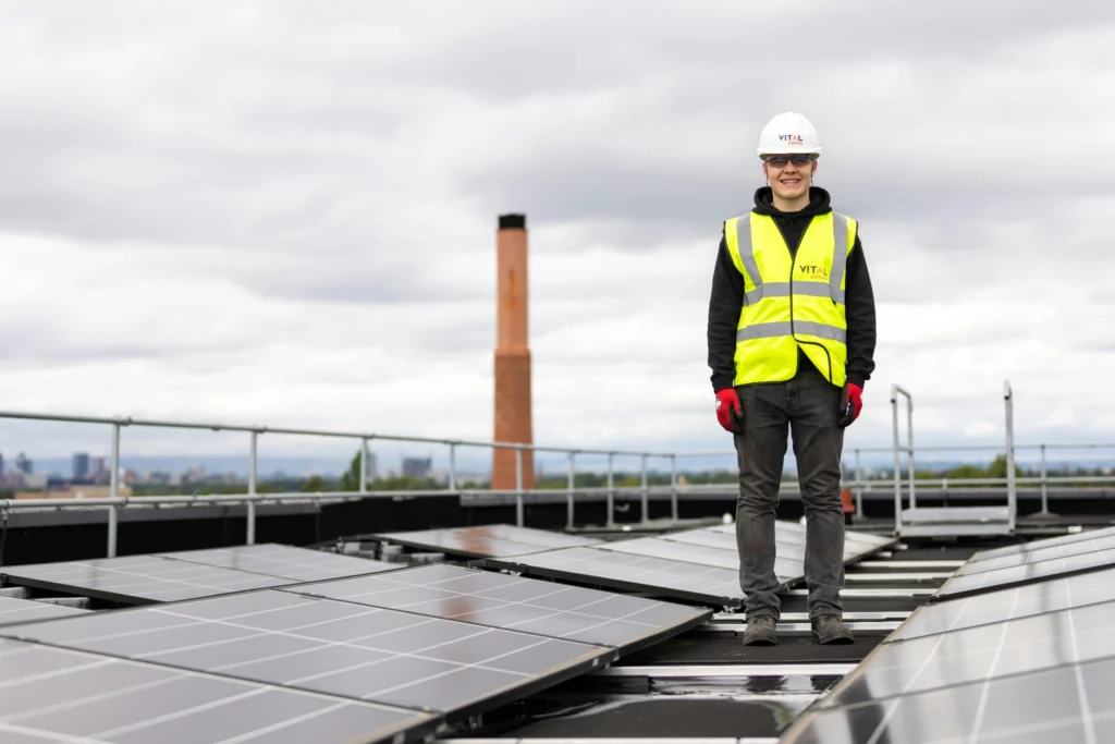 Technicien en sécurité avec casque et gilet réfléchissant debout sur un toit équipé de panneaux solaires.