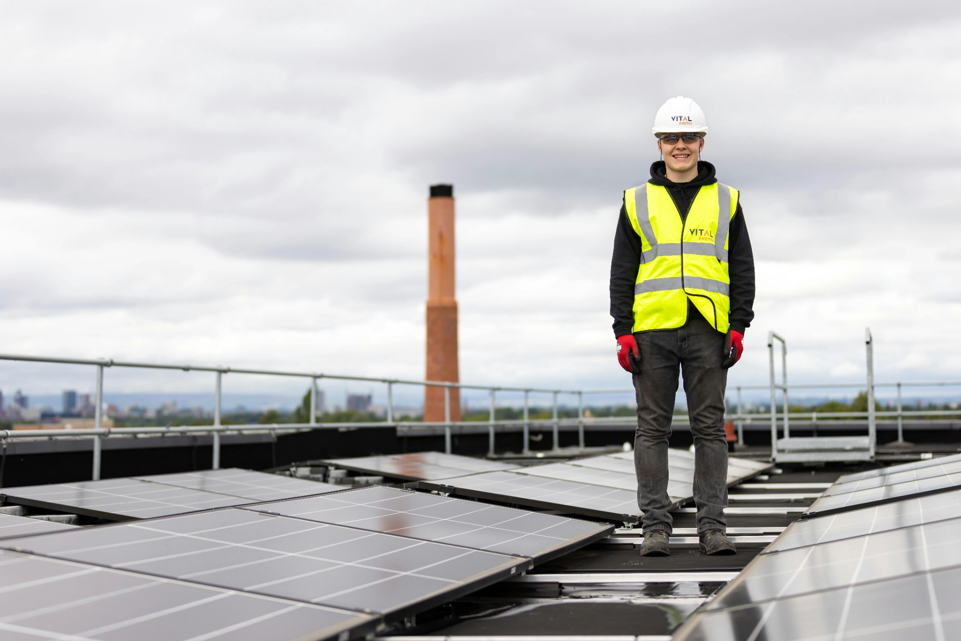 Technicien en sécurité avec casque et gilet réfléchissant debout sur un toit équipé de panneaux solaires.