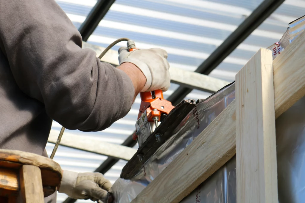 Ouvrier effectuant des travaux de toiture - RenovSmart Un ouvrier utilise un pistolet à air comprimé pour fixer un revêtement sur une structure en bois pendant des travaux de toiture.
