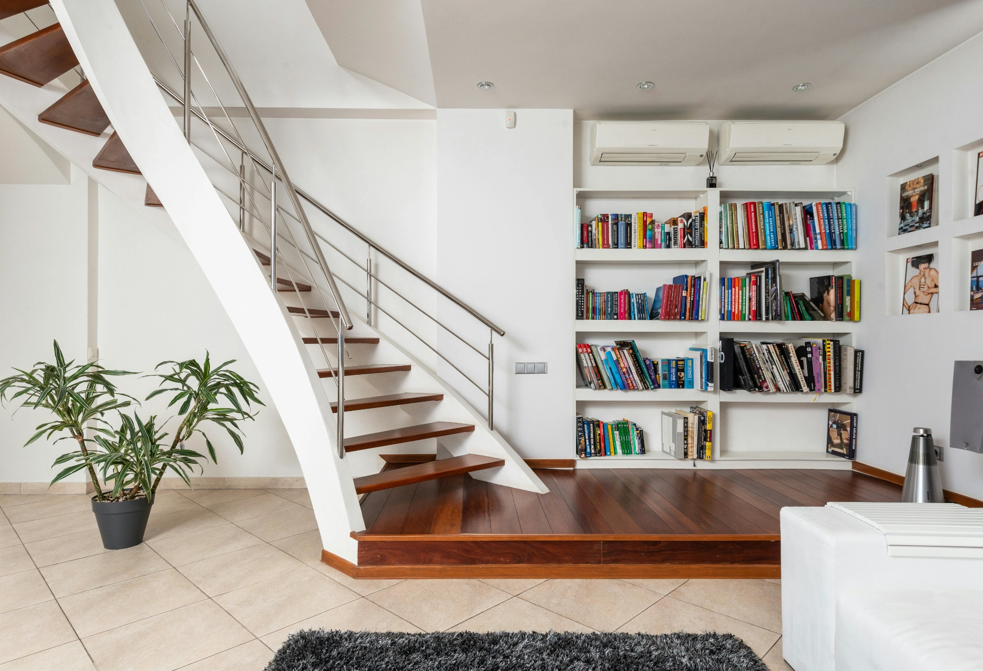 Intérieur moderne avec un escalier en bois, une étagère remplie de livres et des plantes décoratives.
