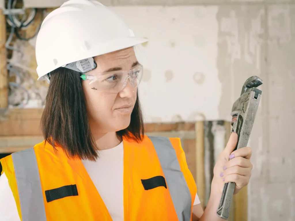 Une professionnelle de la construction, portant un casque blanc et des lunettes de protection, examine une clé à molette avec une expression perplexe.