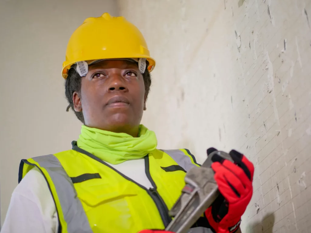 Une professionnelle portant un casque jaune, un gilet de sécurité et des gants rouges tient une clé à molette, en regardant attentivement un mur sur un chantier de construction.