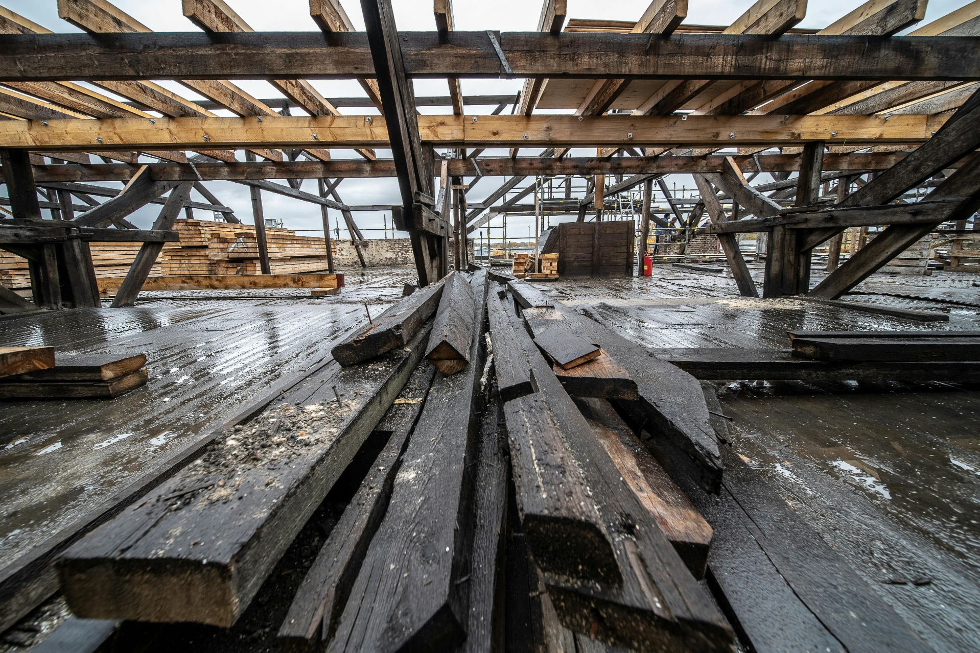 Vue d'un chantier de construction avec des poutres en bois disposées sur le sol, sous une structure de toiture en construction.