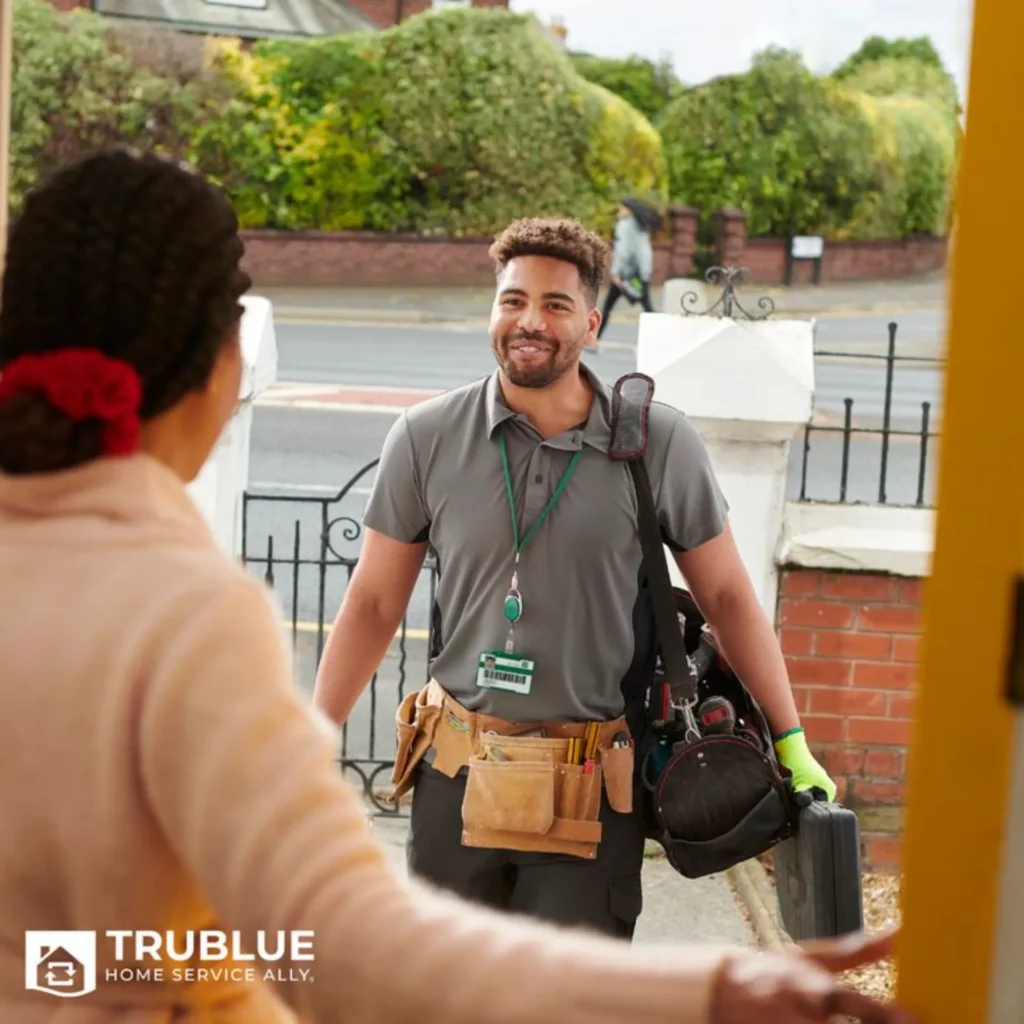 Un plombier en uniforme, portant une ceinture à outils et un sac à dos, sourit tout en entrant chez un client pour un service à domicile.