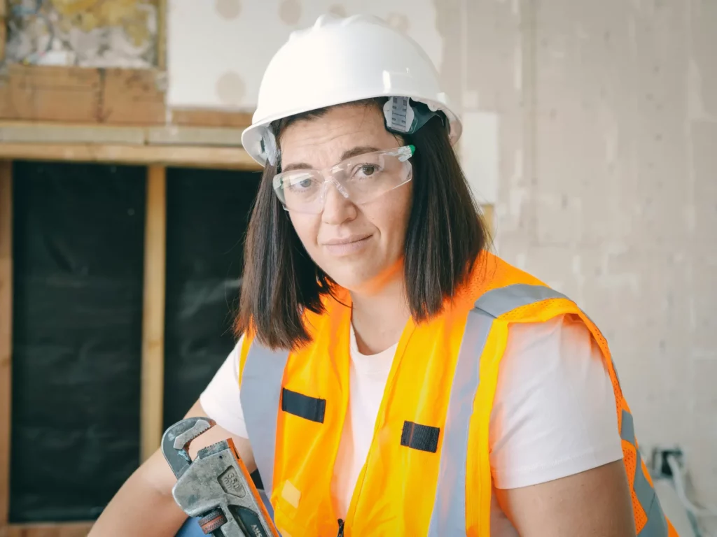 Une femme plombier, portant un casque blanc et des lunettes de sécurité, tient une clé à molette dans un chantier de construction.