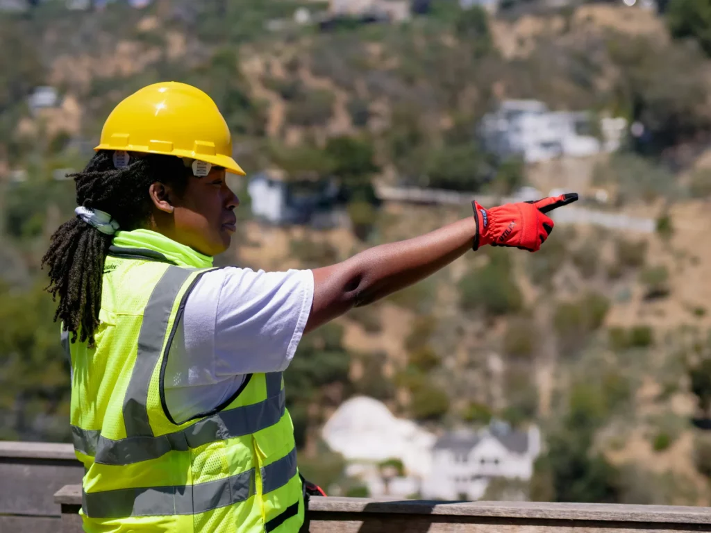 Une professionnelle de la construction, portant un casque jaune et un gilet de sécurité, pointe un objet au loin sur un chantier en extérieur.