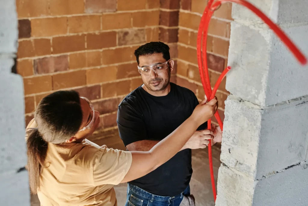 Deux travailleurs en chantier manipulent des tuyaux rouges pour une installation, en discutant des détails de leur tâche.